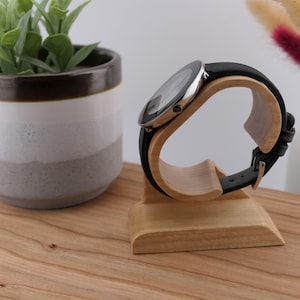 May include: A black leather strap watch with a silver case sits on a light-coloured wooden watch stand. The stand is curved to hold the watch. A small potted plant and vase are in the background.