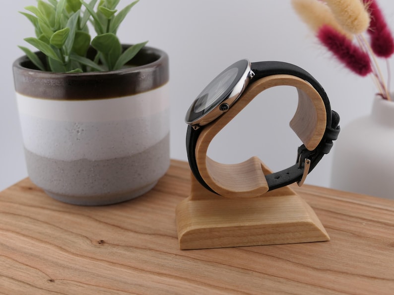 May include: A black leather strap watch with a silver case sits on a light-coloured wooden watch stand. The stand is curved to hold the watch. A small potted plant and vase are in the background.