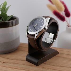 May include: A wristwatch with a dark blue face and silver casing is displayed on a dark brown wooden watch stand. The watch has a brown leather strap. The stand is on a wooden surface, with a small plant and vase in the background.