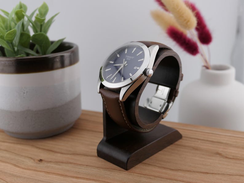 May include: A wristwatch with a dark blue face and silver casing is displayed on a dark brown wooden watch stand. The watch has a brown leather strap. The stand is on a wooden surface, with a small plant and vase in the background.