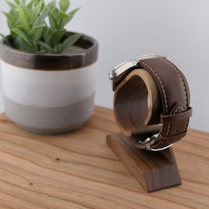 May include: A brown leather watch with a silver case sits on a wooden watch stand. The stand is made of dark wood and has a curved cradle to hold the watch. A small potted plant is in the background on a wooden surface.