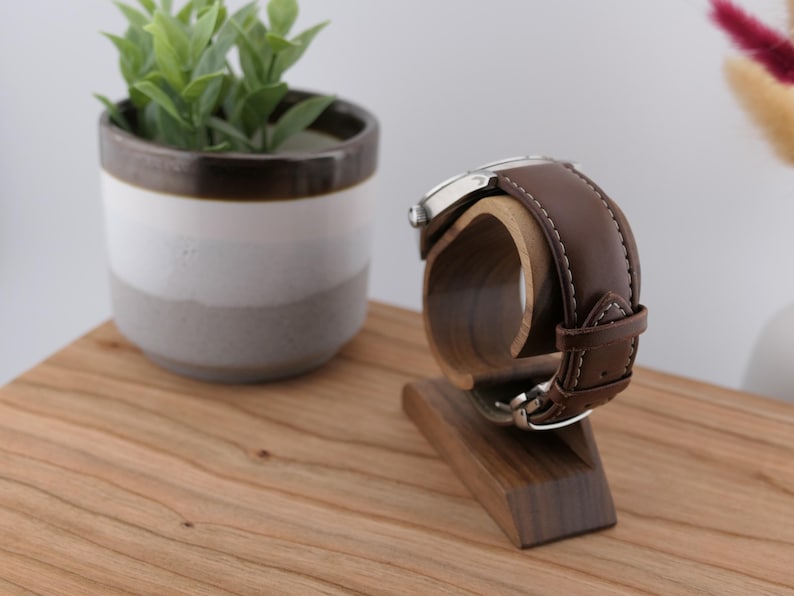 May include: A brown leather watch with a silver case sits on a wooden watch stand. The stand is made of dark wood and has a curved cradle to hold the watch. A small potted plant is in the background on a wooden surface.