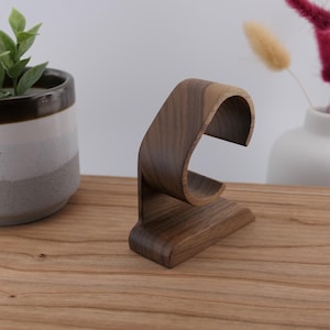 May include: A dark brown wooden watch stand with a curved design. The stand is made of wood and has a base for stability. The watch stand is displayed on a wooden surface, with a potted plant and vase in the background.
