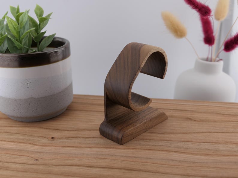 May include: A dark brown wooden watch stand with a curved design. The stand is made of wood and has a base for stability. The watch stand is displayed on a wooden surface, with a potted plant and vase in the background.