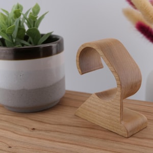 May include: A wooden watch stand with a curved design. The stand is made of light-coloured wood and has a base and a curved top to hold a watch. A small potted plant and a vase with decorative stems are in the background.