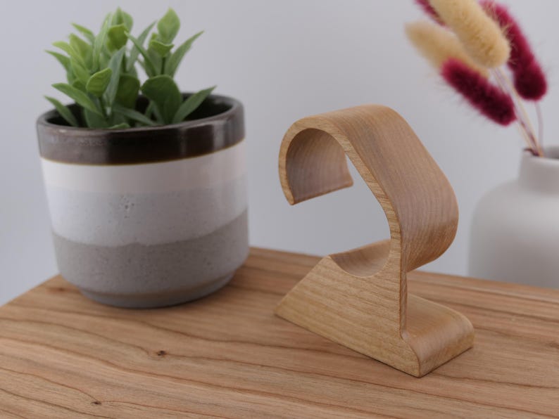 May include: A wooden watch stand with a curved design. The stand is made of light-coloured wood and has a base and a curved top to hold a watch. A small potted plant and a vase with decorative stems are in the background.