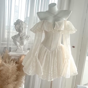May include: A cream-colored lace mini dress featuring an off-the-shoulder neckline with ruffled sleeves, long flared sleeves, a corset-style bodice with a pointed waistline, and thin spaghetti straps that cross over the chest area.