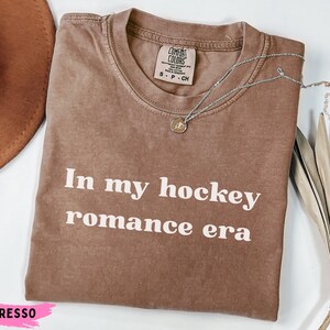 May include: Espresso-coloured t-shirt with the text "In my hockey romance era" in white. A silver necklace with a pendant rests on the shirt. The shirt is folded and displayed with a brown hat and dried foliage.