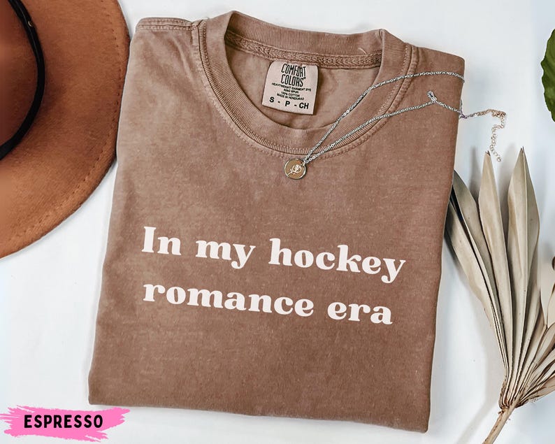 May include: Espresso-coloured t-shirt with the text "In my hockey romance era" in white. A silver necklace with a pendant rests on the shirt. The shirt is folded and displayed with a brown hat and dried foliage.