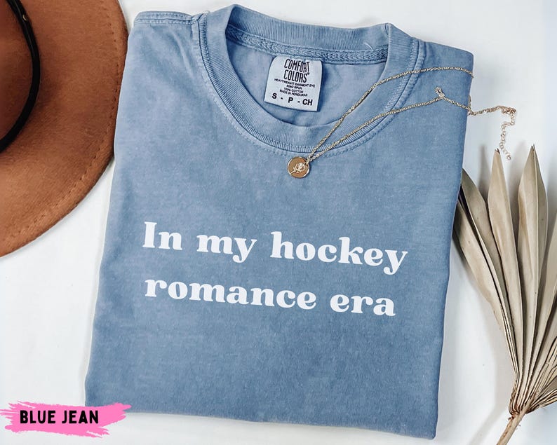 May include: A blue jean coloured t-shirt with the text "In my hockey romance era" in white. A gold necklace with a pendant is draped across the shirt. The shirt is folded on a white surface.