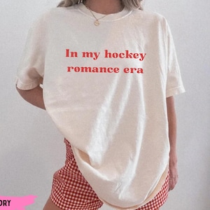 Könnte beinhalten: Cremefarbenes T-Shirt mit dem roten Text "In my hockey romance era". Das Shirt wird mit rot-weiß karierten Shorts getragen. Das Outfit hat einen entspannten, lässigen Stil.
