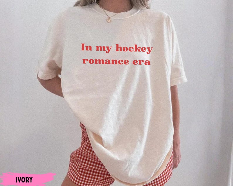 May include: Cream-coloured t-shirt with the red text "In my hockey romance era". The shirt is worn with red and white checked shorts. The outfit has a relaxed, casual style.