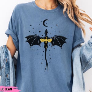 K&ouml;nnte beinhalten: Ein blaues T-Shirt in Jeansoptik mit einem schwarzen Grafikdruck eines Drachen mit gelben Fl&uuml;geln, der unter einer Mondsichel und verstreuten Sternen abgebildet ist. Der Drache ist im Sturzflug mit einem langen, schmalen Schwanz dargestellt.