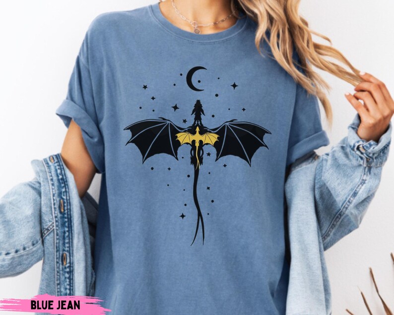 K&ouml;nnte beinhalten: Ein blaues T-Shirt in Jeansoptik mit einem schwarzen Grafikdruck eines Drachen mit gelben Fl&uuml;geln, der unter einer Mondsichel und verstreuten Sternen abgebildet ist. Der Drache ist im Sturzflug mit einem langen, schmalen Schwanz dargestellt.