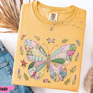 May include: Mustard-coloured t-shirt featuring a patchwork butterfly design. The butterfly wings are made of floral and patterned fabric pieces. The shirt has a crew neck and short sleeves. A silver necklace with a small pendant is draped on the shirt. The text "COMFORT COLORS" is visible on the tag.