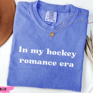 May include: A blue t-shirt with the text "In my hockey romance era" in white. The shirt has a crew neck and short sleeves. A gold necklace with a pendant is draped across the shirt. The shirt is labeled "Flo Blue".