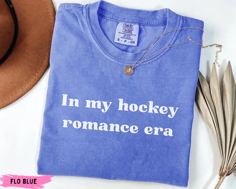 May include: A blue t-shirt with the text "In my hockey romance era" in white. The shirt has a crew neck and short sleeves. A gold necklace with a pendant is draped across the shirt. The shirt is labeled "Flo Blue".