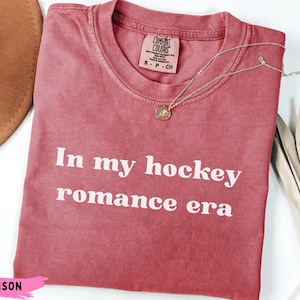 May include: Crimson t-shirt with the white text "In my hockey romance era". The shirt has a crew neck and a small tag. A silver necklace with a pendant is draped across the shirt. A brown hat and dried leaves are in the background.
