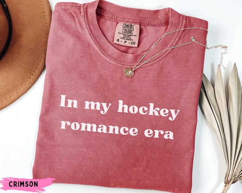 May include: Crimson t-shirt with the white text "In my hockey romance era". The shirt has a crew neck and a small tag. A silver necklace with a pendant is draped across the shirt. A brown hat and dried leaves are in the background.