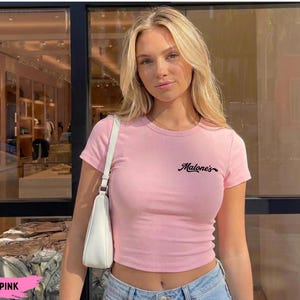 May include: A light pink short-sleeved t-shirt featuring the text "Malone's" in black cursive script on the upper left chest. The shirt has a crew neckline and a fitted silhouette, paired with a white shoulder bag and blue denim jeans.