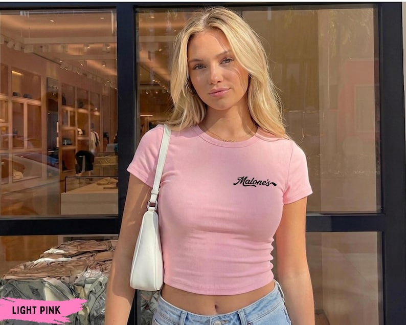 May include: A light pink short-sleeved t-shirt featuring the text "Malone's" in black cursive script on the upper left chest. The shirt has a crew neckline and a fitted silhouette, paired with a white shoulder bag and blue denim jeans.