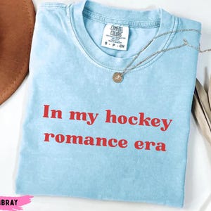 May include: Pale blue t-shirt featuring the red text "In my hockey romance era". The shirt has a crew neck and short sleeves. A silver necklace with a pendant is draped across the shirt.