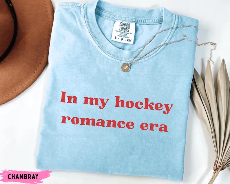 May include: Pale blue t-shirt featuring the red text "In my hockey romance era". The shirt has a crew neck and short sleeves. A silver necklace with a pendant is draped across the shirt.