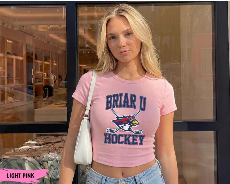 May include: A light pink short-sleeved crop top featuring the text BRIAR U HOCKEY in dark blue letters with a graphic of a bird mascot head and two crossed hockey sticks in the center. The shirt is paired with a white shoulder bag.