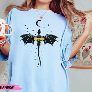 K&ouml;nnte beinhalten: Ein hellblaues T-Shirt mit einer schwarz-gelben Grafik eines Drachen mit ausgebreiteten Fl&uuml;geln unter einer Mondsichel und Sternen. Das Motiv ist mittig auf der Brust des Kleidungsst&uuml;cks platziert, das kurze &Auml;rmel und einen Rundhalsausschnitt hat.