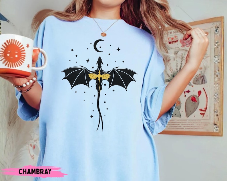 K&ouml;nnte beinhalten: Ein hellblaues T-Shirt mit einer schwarz-gelben Grafik eines Drachen mit ausgebreiteten Fl&uuml;geln unter einer Mondsichel und Sternen. Das Motiv ist mittig auf der Brust des Kleidungsst&uuml;cks platziert, das kurze &Auml;rmel und einen Rundhalsausschnitt hat.