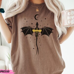 K&ouml;nnte beinhalten: Ein braunes Kurzarm-T-Shirt mit einer schwarz-gelben Grafik eines Drachen mit ausgebreiteten Fl&uuml;geln unter einer Mondsichel und kleinen Sternen. Das T-Shirt hat einen Rundhalsausschnitt, eine lockere Passform und das Wort ESPRESSO unten.