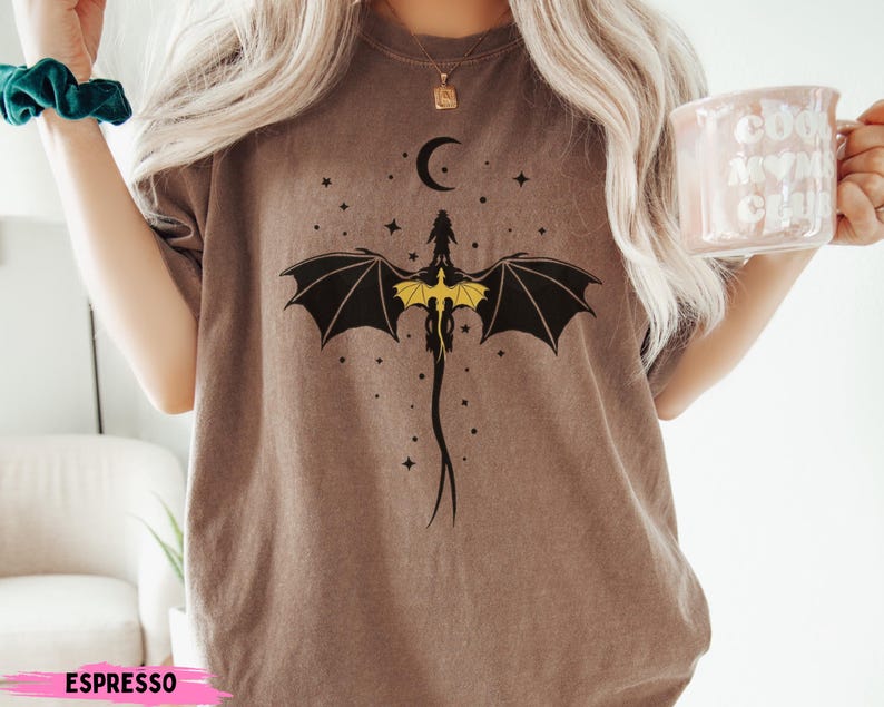 K&ouml;nnte beinhalten: Ein braunes Kurzarm-T-Shirt mit einer schwarz-gelben Grafik eines Drachen mit ausgebreiteten Fl&uuml;geln unter einer Mondsichel und kleinen Sternen. Das T-Shirt hat einen Rundhalsausschnitt, eine lockere Passform und das Wort ESPRESSO unten.
