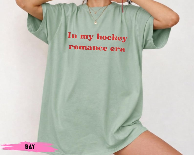 May include: A pale green t-shirt featuring the red text "In my hockey romance era". The shirt has a loose fit and short sleeves. The word "BAY" is written in pink at the bottom.
