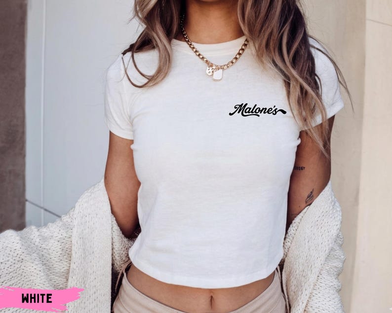 May include: A white short-sleeved t-shirt featuring the word Malone's printed in black cursive script on the upper left chest area. The shirt has a crew neckline and a fitted silhouette, paired with a gold chain necklace and a cream-colored cardigan.