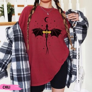 K&ouml;nnte beinhalten: Ein dunkelrotes T-Shirt mit Rundhalsausschnitt und einer schwarz-gelben Grafik eines Drachen mit ausgebreiteten Fl&uuml;geln unter einer Mondsichel und Sternen. Das Shirt wird mit einem schwarz-wei&szlig; karierten Hemd und schwarzen Shorts getragen.