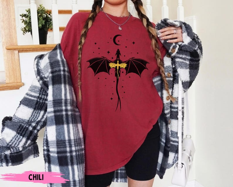 K&ouml;nnte beinhalten: Ein dunkelrotes T-Shirt mit Rundhalsausschnitt und einer schwarz-gelben Grafik eines Drachen mit ausgebreiteten Fl&uuml;geln unter einer Mondsichel und Sternen. Das Shirt wird mit einem schwarz-wei&szlig; karierten Hemd und schwarzen Shorts getragen.
