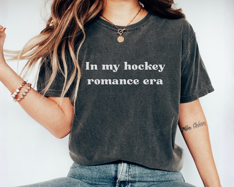 May include: Dark grey t-shirt with the white text "In my hockey romance era". The shirt has a washed-out, vintage appearance. The model is wearing blue jeans and a gold necklace.