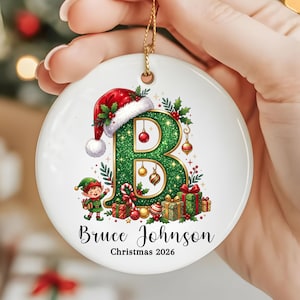 May include: A round white ceramic Christmas ornament featuring a large green glittery letter B decorated with a Santa hat, holly, ornaments, and a small elf. Below the letter, the text reads Bruce Johnson and Christmas 2026.