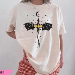 K&ouml;nnte beinhalten: Ein elfenbeinfarbenes Kurzarm-T-Shirt mit einer schwarz-gelben Grafik eines Drachen mit ausgebreiteten Fl&uuml;geln, der unter einer Mondsichel platziert und von kleinen Sternen umgeben ist. Das Motiv ist mittig auf der Brust platziert.