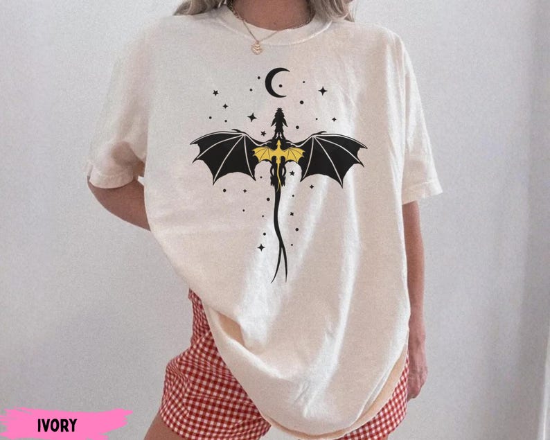 K&ouml;nnte beinhalten: Ein elfenbeinfarbenes Kurzarm-T-Shirt mit einer schwarz-gelben Grafik eines Drachen mit ausgebreiteten Fl&uuml;geln, der unter einer Mondsichel platziert und von kleinen Sternen umgeben ist. Das Motiv ist mittig auf der Brust platziert.