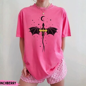 K&ouml;nnte beinhalten: Ein leuchtend pinkfarbenes T-Shirt mit kurzen &Auml;rmeln und einer schwarzen Grafik eines Drachen mit gelben Fl&uuml;geln unter einem Halbmond und Sternen. Das T-Shirt wird mit einer rosa-wei&szlig; karierten Shorts kombiniert. Der Text CRUNCHBERRY ist sichtbar.