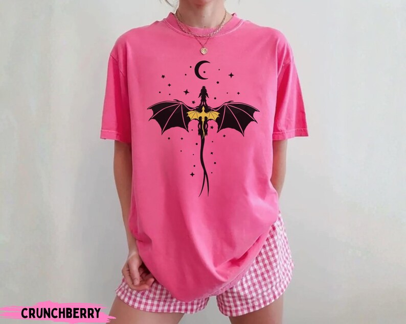 K&ouml;nnte beinhalten: Ein leuchtend pinkfarbenes T-Shirt mit kurzen &Auml;rmeln und einer schwarzen Grafik eines Drachen mit gelben Fl&uuml;geln unter einem Halbmond und Sternen. Das T-Shirt wird mit einer rosa-wei&szlig; karierten Shorts kombiniert. Der Text CRUNCHBERRY ist sichtbar.