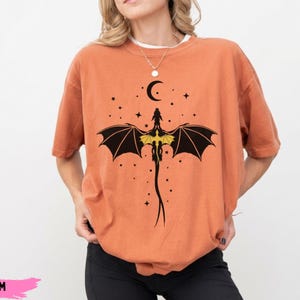 K&ouml;nnte beinhalten: Ein &uuml;bergro&szlig;es, rostorangefarbenes T-Shirt mit einem schwarzen Grafikdruck eines Drachen mit ausgebreiteten Fl&uuml;geln, einem gelben Brustdetail sowie einem Halbmond und kleinen Sternen. Das T-Shirt hat kurze &Auml;rmel und einen Rundhalsausschnitt.
