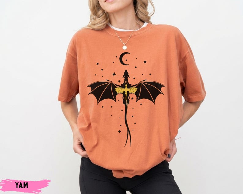 K&ouml;nnte beinhalten: Ein &uuml;bergro&szlig;es, rostorangefarbenes T-Shirt mit einem schwarzen Grafikdruck eines Drachen mit ausgebreiteten Fl&uuml;geln, einem gelben Brustdetail sowie einem Halbmond und kleinen Sternen. Das T-Shirt hat kurze &Auml;rmel und einen Rundhalsausschnitt.