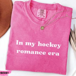 May include: A vibrant pink t-shirt with the white text "In my hockey romance era". The shirt features a crew neck and short sleeves. A gold necklace with a pendant is placed on the shirt.