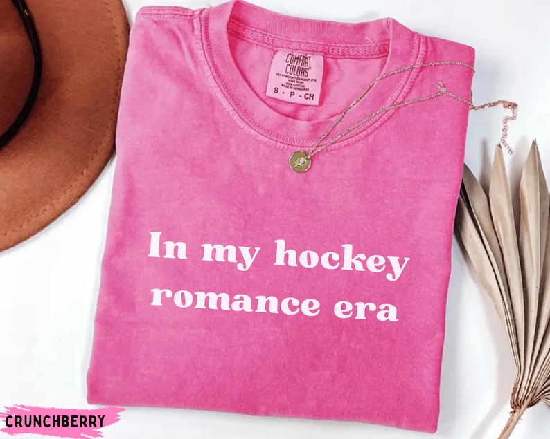 May include: A vibrant pink t-shirt with the white text "In my hockey romance era". The shirt features a crew neck and short sleeves. A gold necklace with a pendant is placed on the shirt.