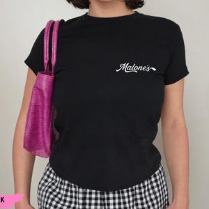 May include: A black short-sleeved t-shirt featuring the white cursive text Malone's on the upper left chest, paired with a bright pink crocodile-textured handbag and black and white checkered bottoms.