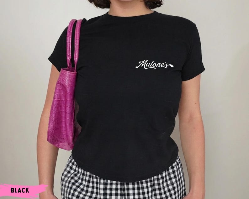 May include: A black short-sleeved t-shirt featuring the white cursive text Malone's on the upper left chest, paired with a bright pink crocodile-textured handbag and black and white checkered bottoms.