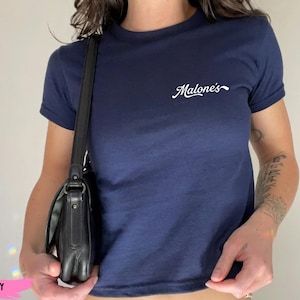 Puede incluir: Una camiseta azul marino de manga corta con la palabra Malone's impresa en letra cursiva blanca en la parte superior izquierda del pecho. La camiseta tiene cuello redondo y un diseño sencillo de color sólido en la parte delantera y trasera.