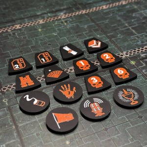 Puede incluir: Una colecci&oacute;n de fichas de juego negras con ilustraciones y n&uacute;meros naranjas. Las fichas presentan varios s&iacute;mbolos, incluyendo calaveras, una huella de mano, una bandera y un micr&oacute;fono. Las fichas est&aacute;n dispuestas sobre una superficie texturizada de color verde oscuro.